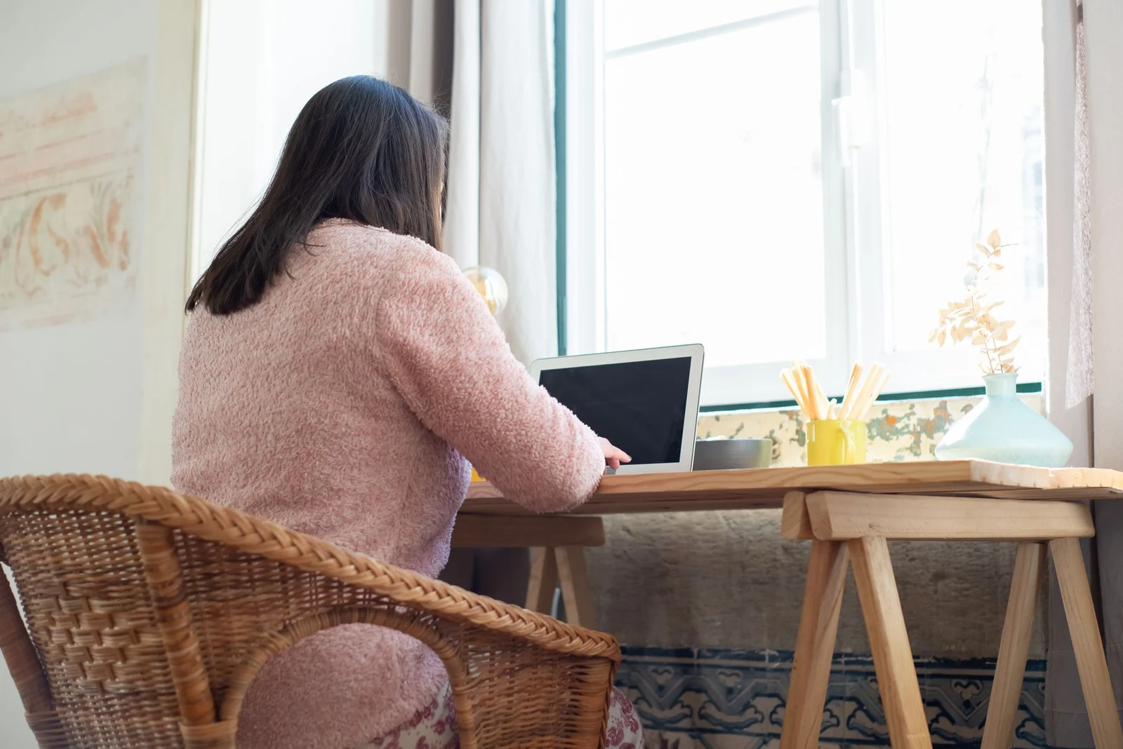 A woman using a laptop at a desk beside a window
